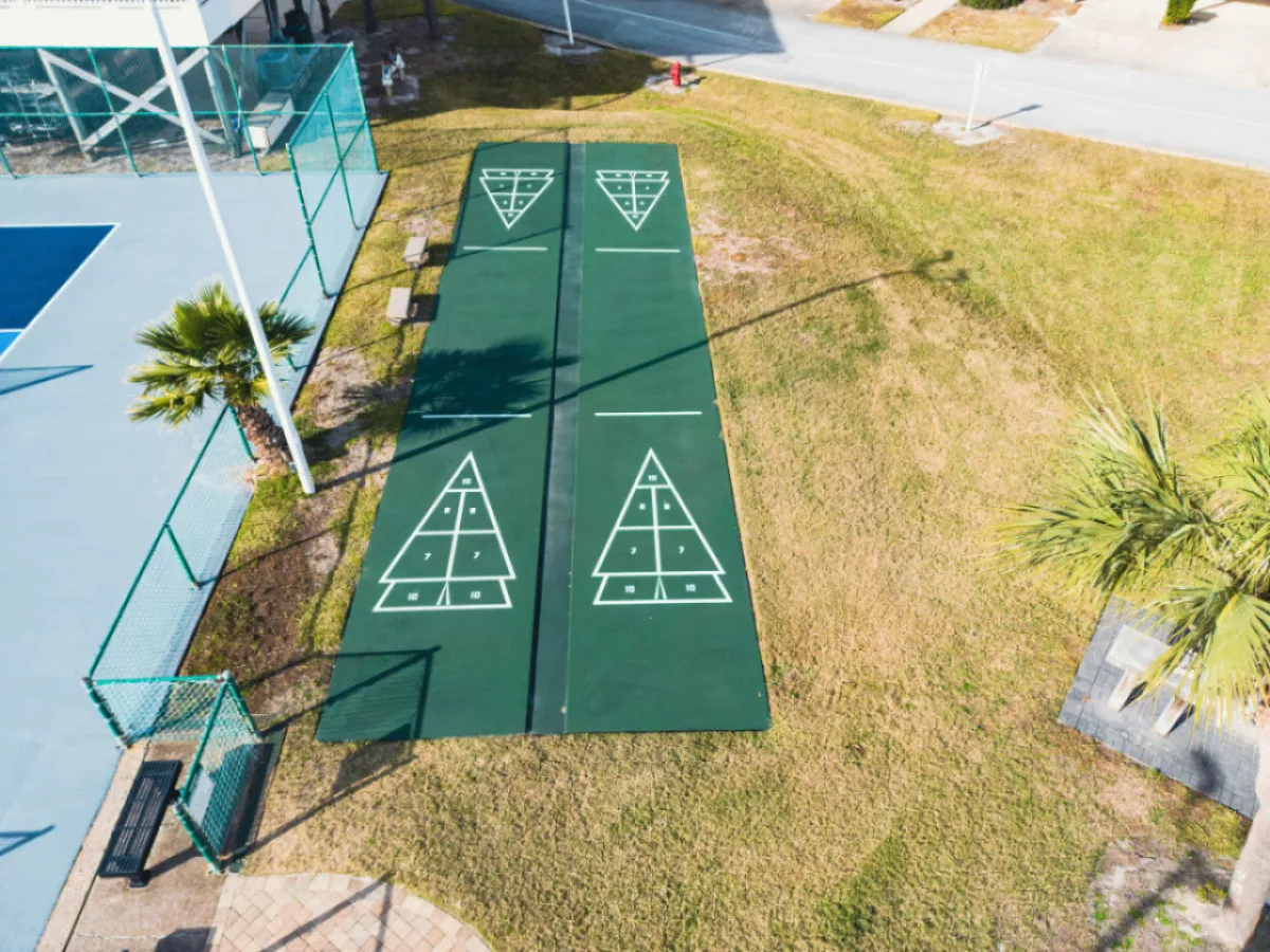 Shuffleboard courts are set amidst surrounding greenery and benches, with a tennis court visible in the background. The environment is open and community-oriented, inviting outdoor recreation and social gatherings.