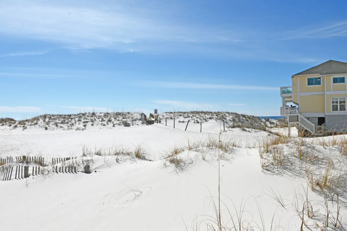 A scenic exterior shot featuring white sand dunes, beach grass, and a view of the ocean with a distinctive yellow house in the frame. The tranquil setting invites relaxation and connection to the coastal environment, hinting at the proximity to the beach.