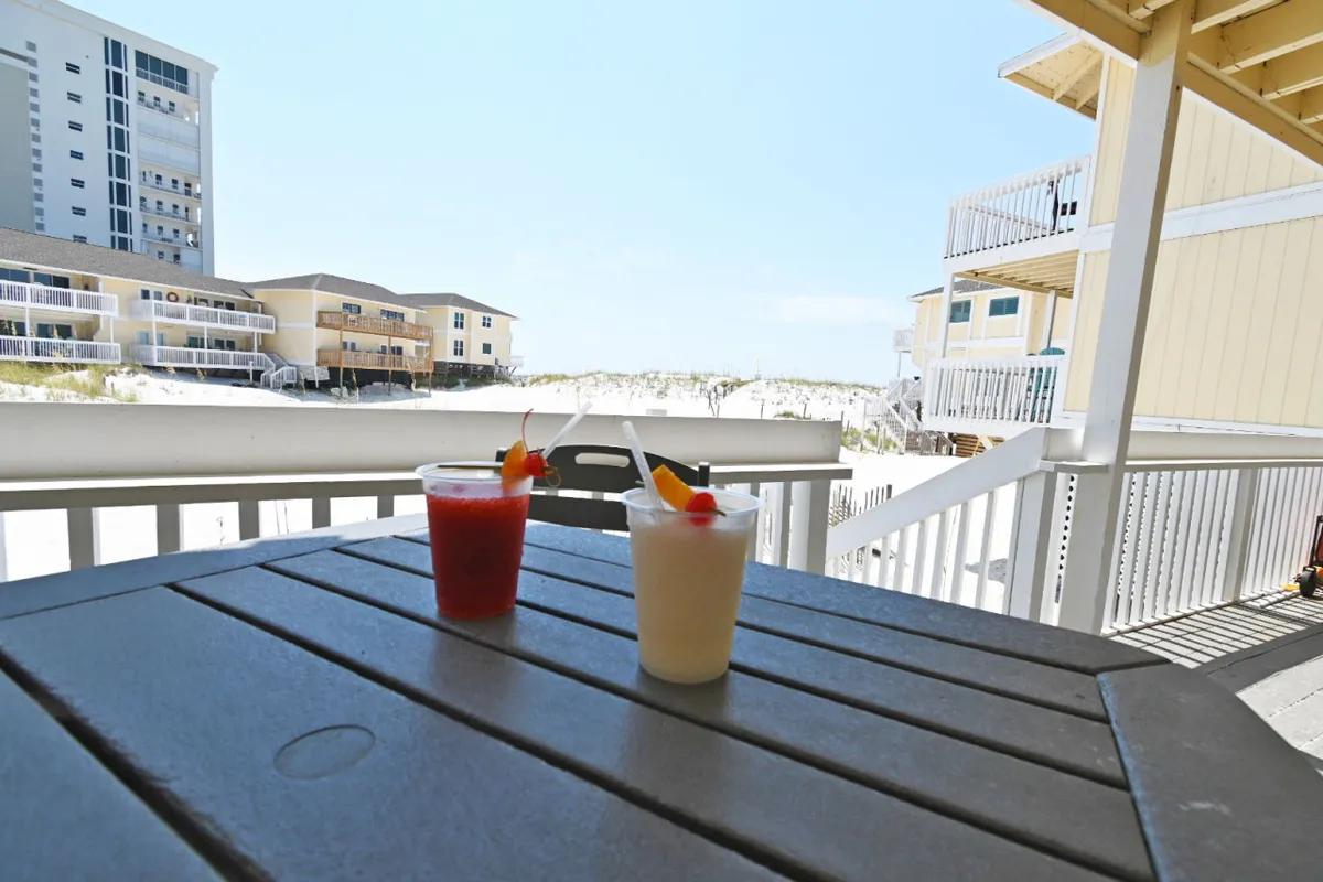 A pair of cocktails rest on a table overlooking a sandy beach, with buildings lining the distant shore. The golden hour lighting hints at a relaxing, romantic ambiance.