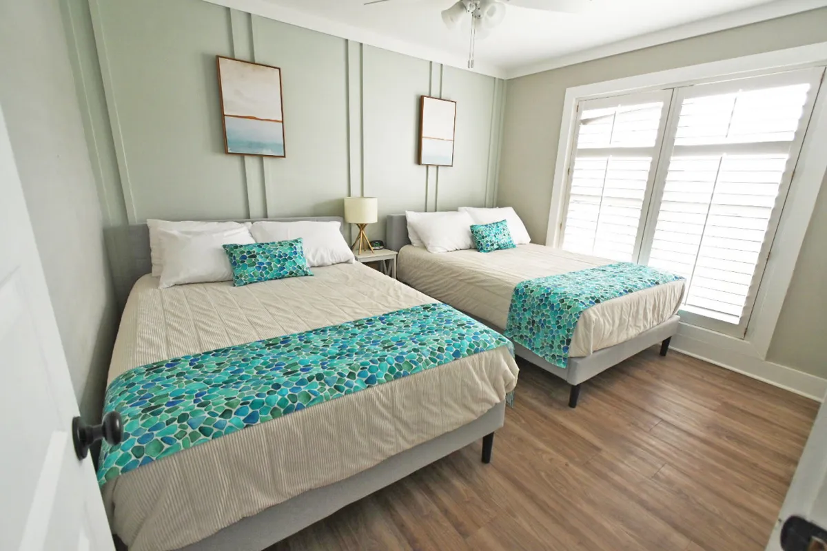 A guest bedroom with two inviting beds dressed in decorative linens and pillows, framed artwork above the headboards, and a sunlit window that fills the room with a comforting glow. The overall mood is warm and welcoming, ideal for families or friends traveling together.