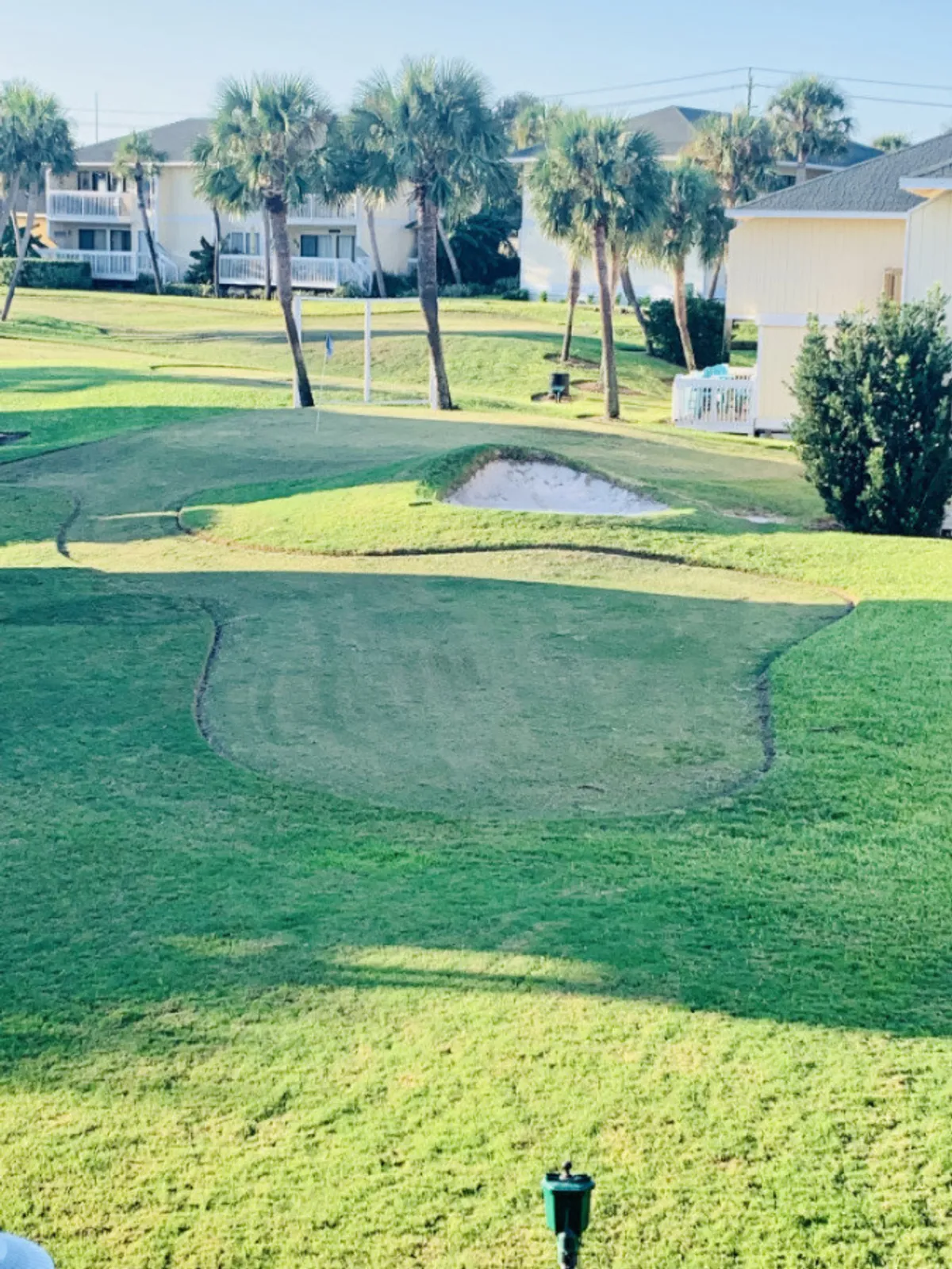 A meticulously maintained Par-3 golf green framed by palm trees, a sand trap, and nearby residential buildings, inviting residents and guests to casual games or afternoon rounds.