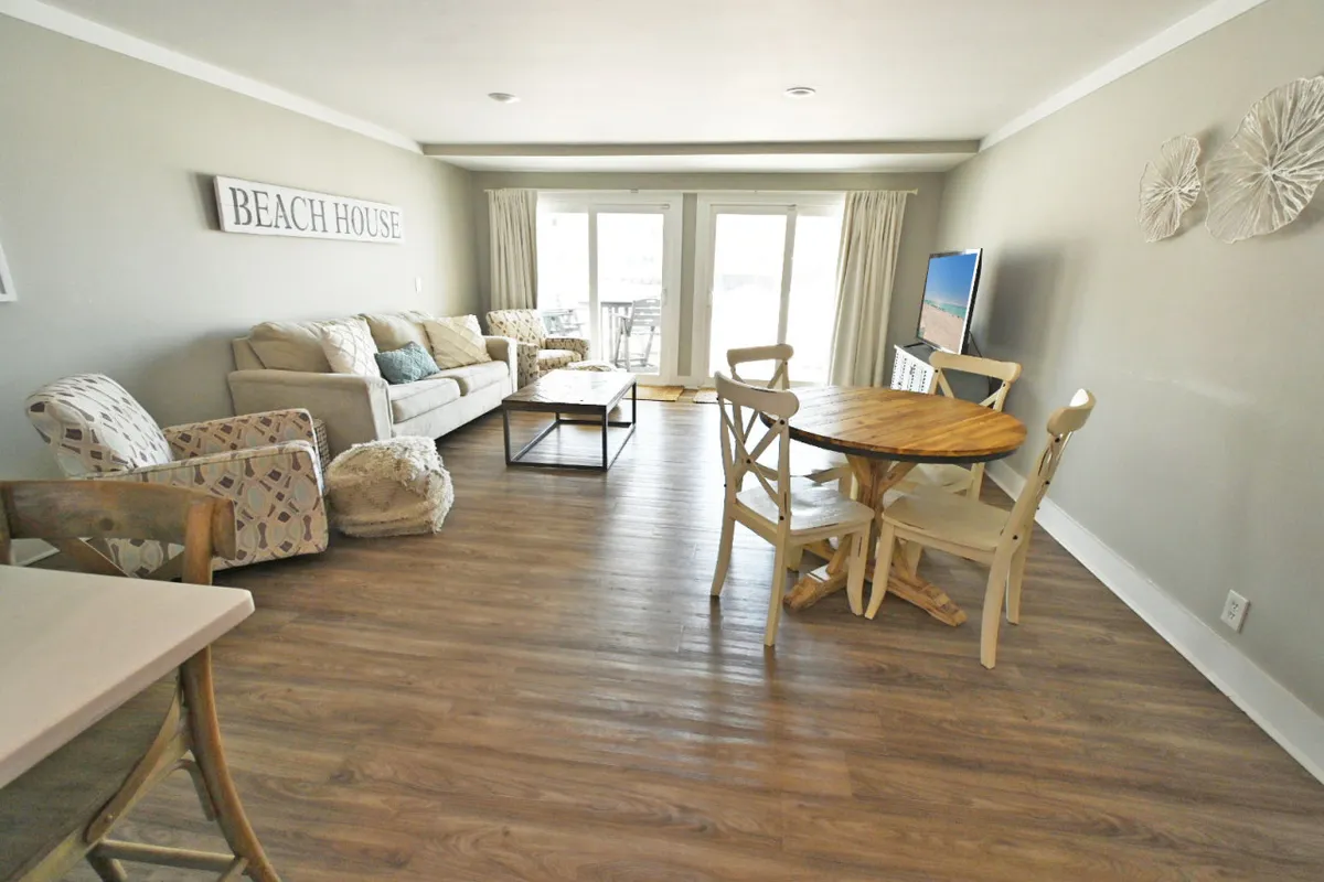 A stylish, beachy living-dining area with a comfortable sofa, dining table, TV, sliding doors letting in gentle sunlight, and decorative wall art. The room blends function and style—ideal for cozy movie nights, shared meals, or simply unwinding after a beach day.