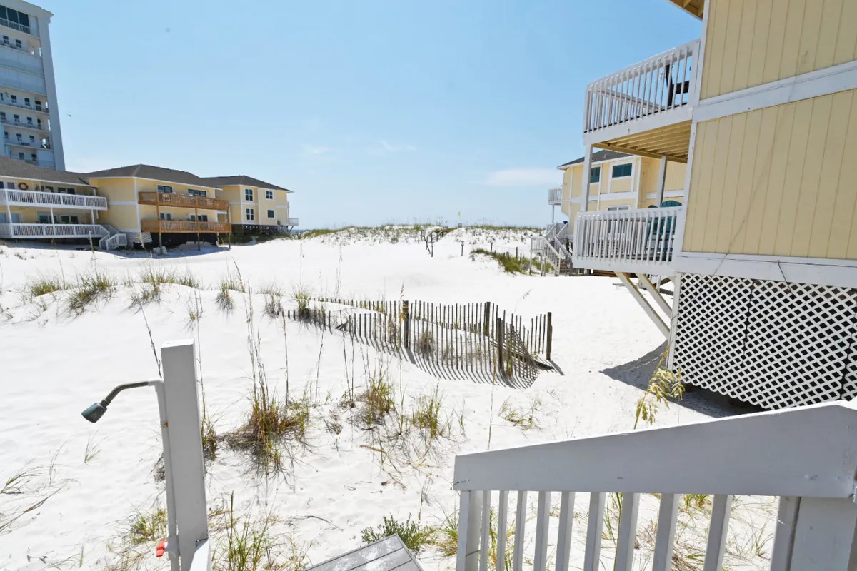 A wooden staircase leads over sand dunes directly to the beach, with the ocean and surrounding residential buildings in the distance. The setting is tranquil and emphasizes easy, direct access to the pristine sands.