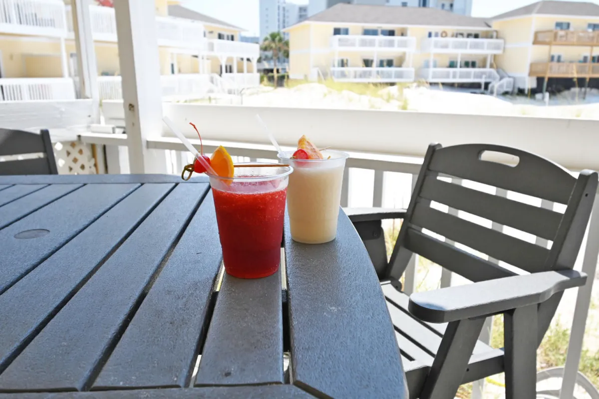 This exterior scene features a table set with colorful cocktails, with background buildings softly lit by the evening sun. The setup suggests a cheerful, social atmosphere perfect for enjoying sunset drinks outdoors.