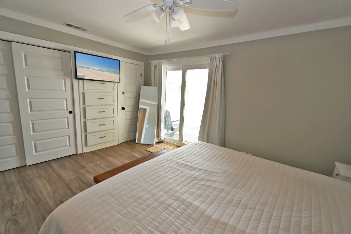 A serene bedroom featuring a king-size bed with decorative pillows, a dresser, a wall-mounted television, and sliding glass doors that allow shimmering natural light to flood the space. The room exudes comfort and offers a direct view outside, hinting at emerald waters beyond. A mirror above the dresser and subtle coastal décor complete the tranquil atmosphere.