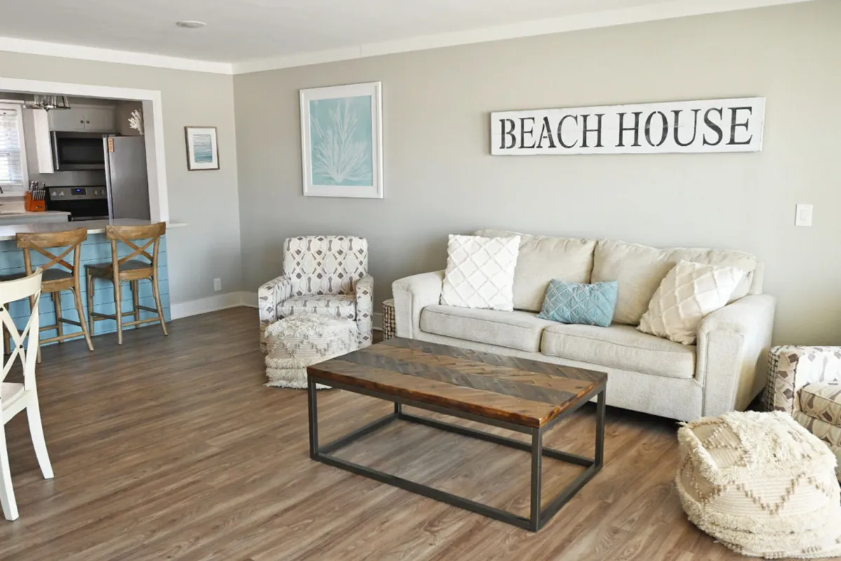 The hero image showcases a sunlit, open-concept living area with a beige sofa adorned with patterned pillows, a charming 'BEACH HOUSE' sign on the wall, a stylish wooden coffee table, and high stools lined up at the kitchen counter. The space is decorated in soothing coastal tones, creating a welcoming and restful atmosphere.