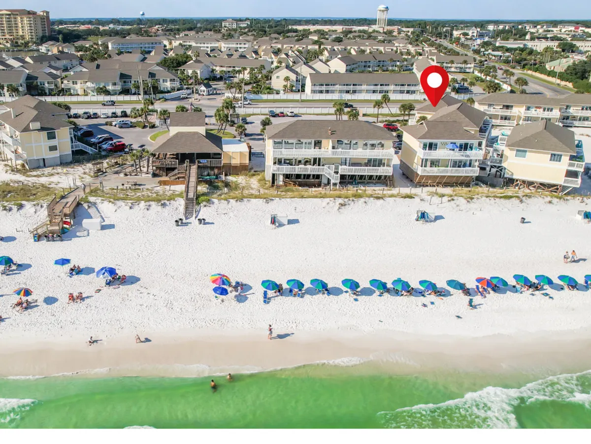 This image presents a vibrant stretch of the Emerald Coast with pristine white sandy beach, colorful umbrellas, emerald green ocean waters, and beachfront condos with direct beach access. The red location marker highlights a specific unit, anchoring the viewer in the exact location for their stay.