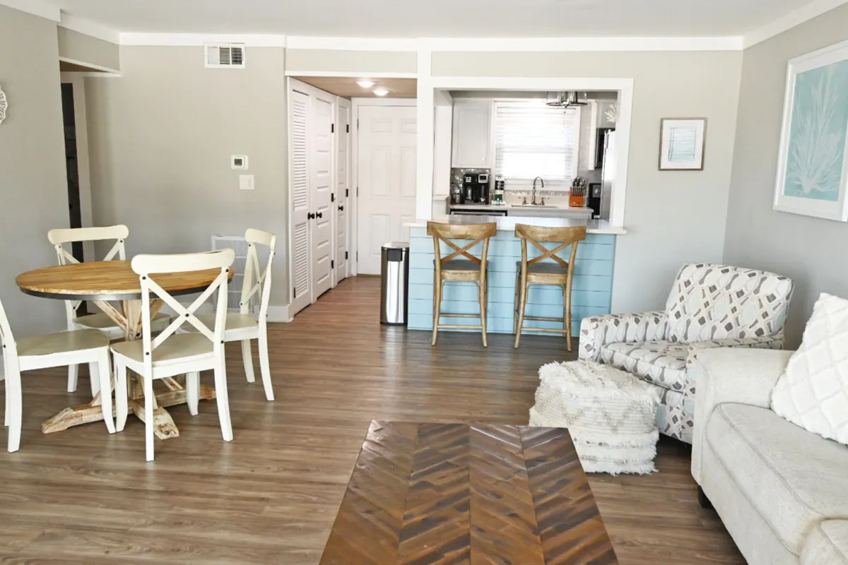 This scene captures the round wooden dining table with white chairs adjacent to a light blue kitchen island, and a comfortable living area with a patterned armchair and neutral sofa. A large window fills the space with natural light and highlights the contemporary, beach-inspired furnishings.