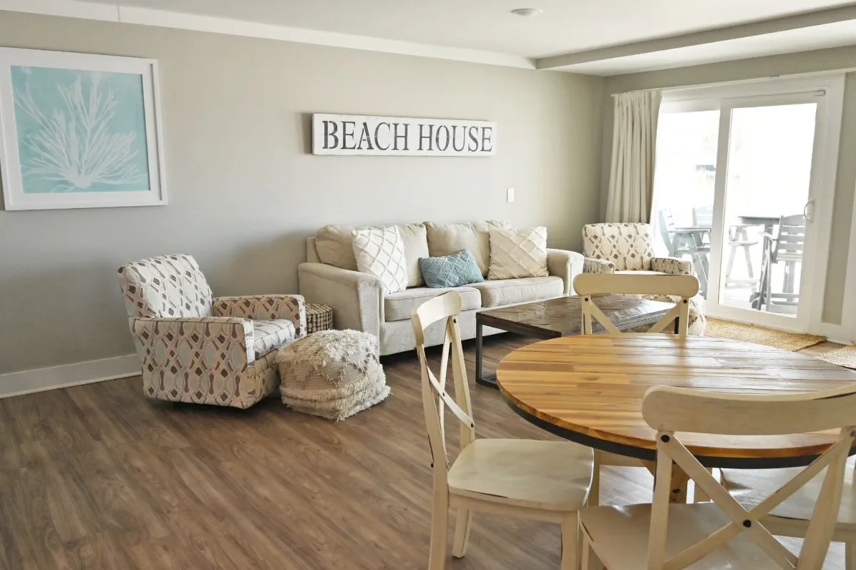 This image highlights a spacious, open-concept living and dining area with a comfortable beige sofa, two patterned armchairs, and a round wooden dining table with chairs. The sliding glass door lets in abundant natural light and leads out to a balcony, reinforcing a seamless indoor-outdoor flow. The décor features subtle coastal touches and a calming, neutral palette.