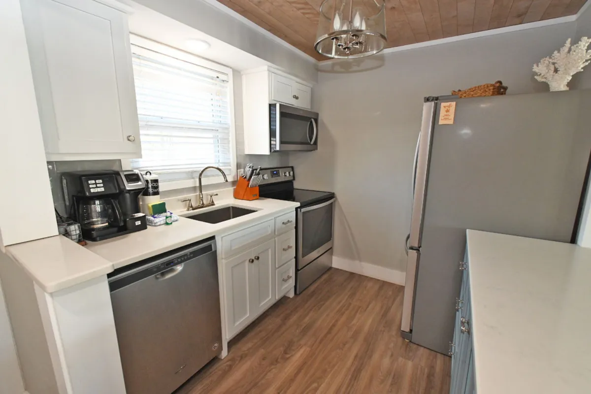 A bright, fully equipped kitchen featuring full-size stainless steel appliances including a refrigerator, stove, microwave, and dishwasher. White cabinetry with granite countertops is complemented by a large window over the sink, allowing natural light to flood the space. Coffee makers and various kitchen tools are neatly arranged on the counters, indicating the kitchen is well-stocked with essentials.