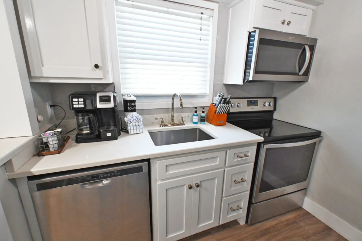 A modern kitchen with stainless steel appliances including a dishwasher, stove, and microwave. The space boasts white cabinetry with sleek hardware, a dedicated coffee station with coffee maker and pods, and a sink area with a window and blinds, contributing to a practical yet inviting atmosphere.
