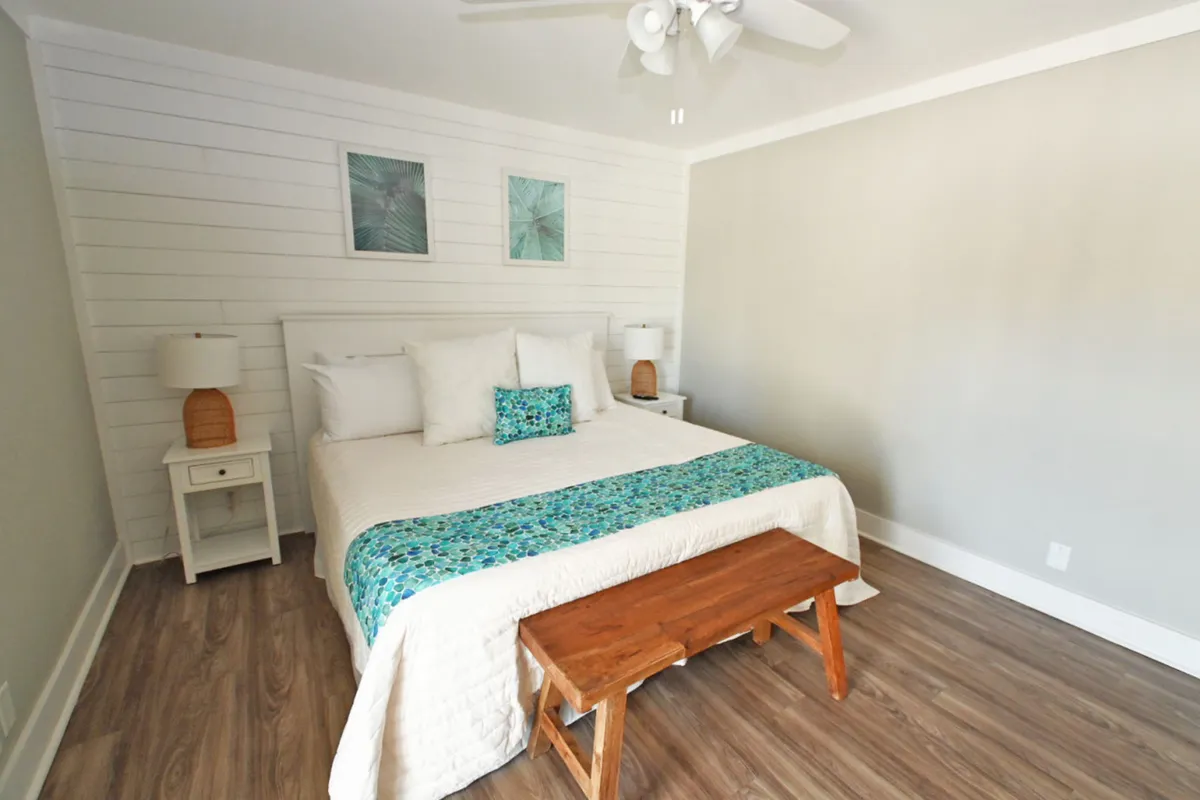 A serene primary bedroom featuring a king-size bed with white and turquoise bedding, a wooden bench at the foot, matching bedside tables with lamps, and two framed tropical leaf artworks above the bed. The space feels airy and restful, embodying a tranquil coastal retreat.
