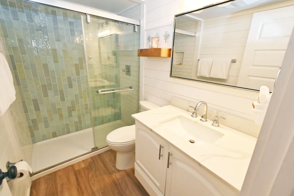 A modern, spa-like bathroom featuring a glass-enclosed shower with green-blue tile, a white vanity with marble countertop and sleek faucet, a large mirror, and a wooden shelf with decorative accents. The space feels bright and rejuvenating.