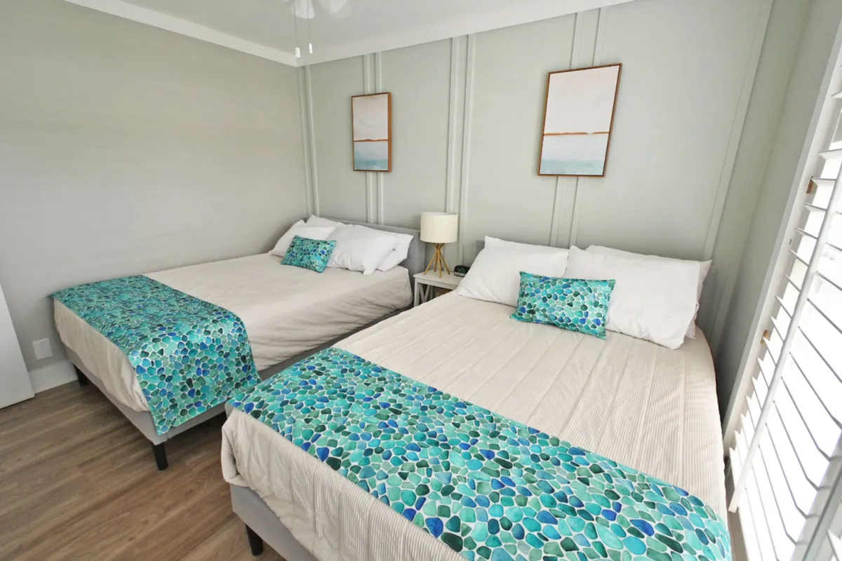 A bright and welcoming guest bedroom with two queen beds, coordinated blue-green patterned runners and pillows, white paneled walls, coastal artwork above each bed, and a window with plantation shutters allowing in natural light. The room is designed for comfort and style.