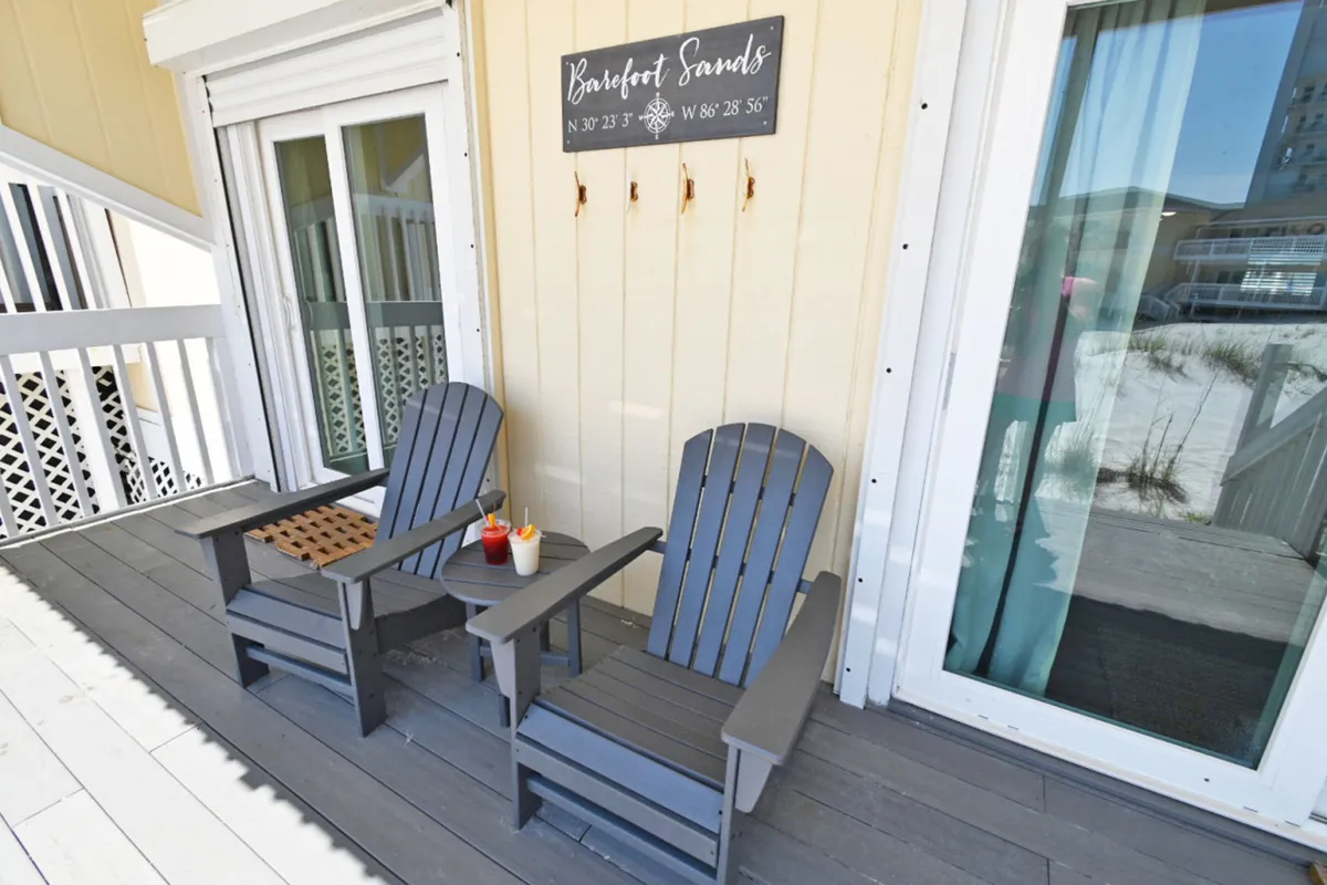 This exterior image shows two gray Adirondack chairs and a small round table with drinks placed on a private deck. The 'Barefoot Sands' sign with coordinates and sliding glass door with light curtains behind it create a cozy, welcoming vibe. The scene suggests a tranquil, private space to relax outdoors and seamlessly connects the interior of the condo to the deck and, by extension, the beach.