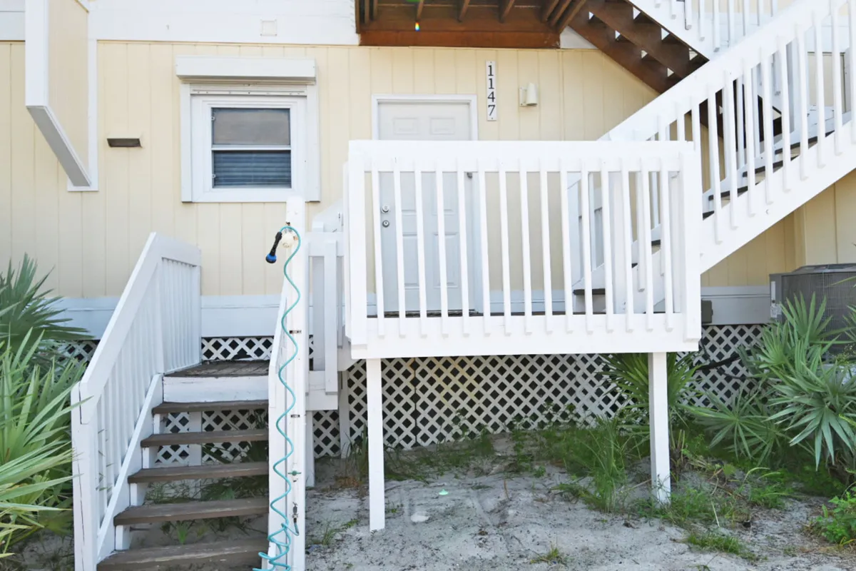 This exterior shot captures the front door labeled 1147, a white wooden staircase, an outdoor shower, and sandy ground with sparse vegetation, embodying a relaxed, beachside retreat vibe and ground-floor convenience.