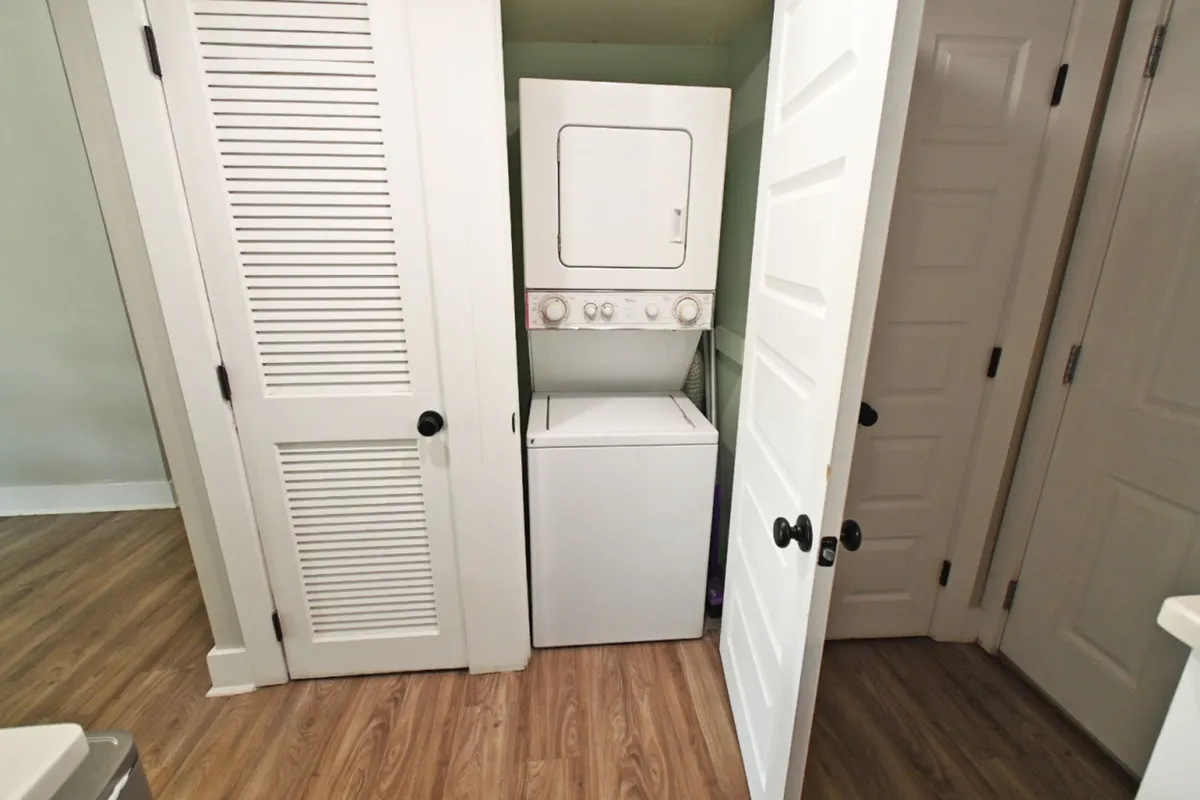 A compact laundry area featuring a stacked washer and dryer unit behind open white doors. The wood-look flooring and neutral wall colors create a calm, homey vibe, emphasizing the convenience of in-unit laundry facilities.