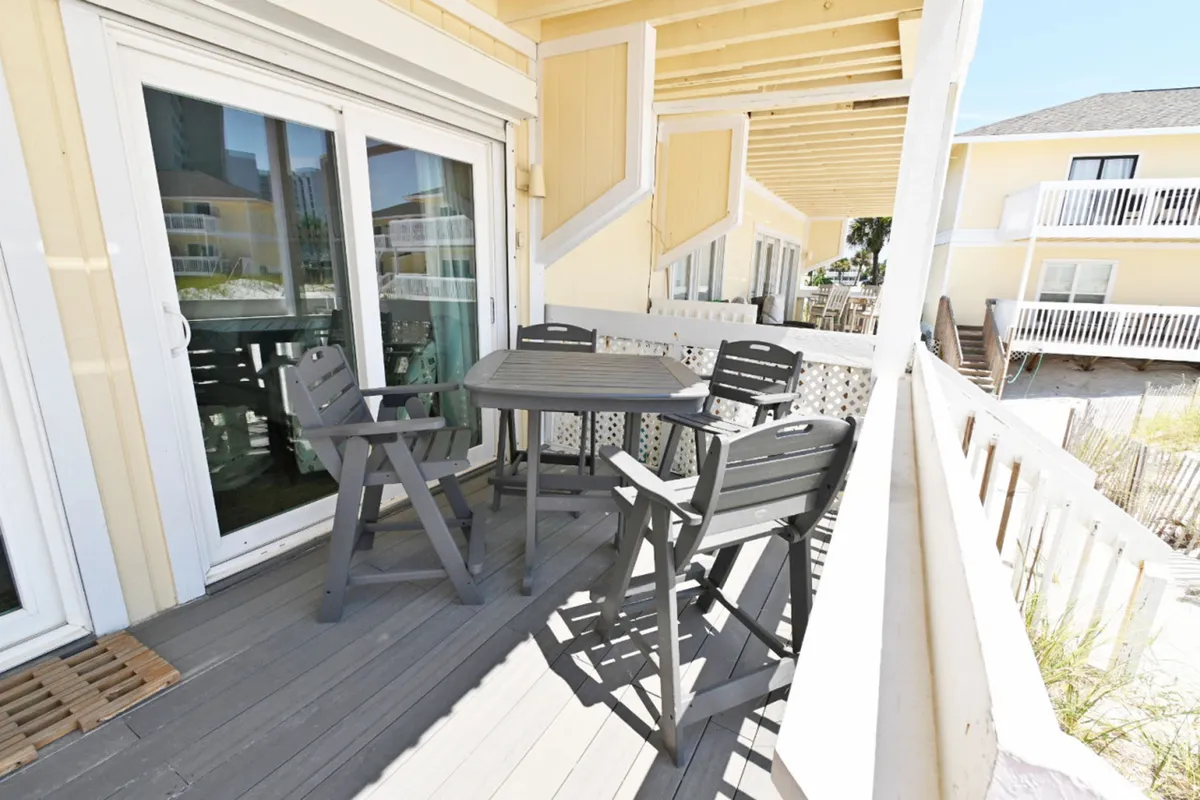This image captures an outdoor dining table with four chairs positioned on a balcony, with sliding glass doors leading inside. The balcony railing offers a partial view of the sandy beach just beyond, suggesting easy access to the shore and the opportunity for al fresco dining with ocean breezes.