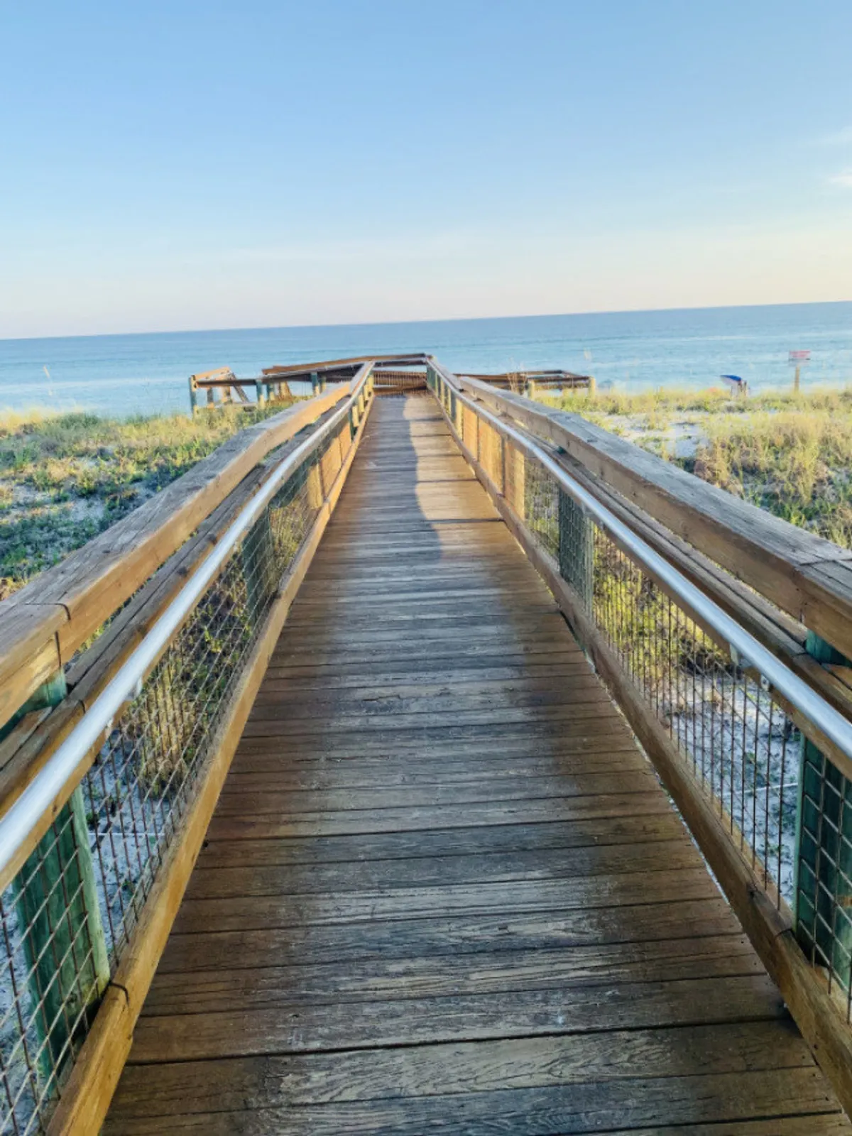 A wooden boardwalk leads through pristine white sand dunes directly to the calm blue ocean waters under a clear blue sky. The boardwalk is inviting and evokes a sense of anticipation for reaching the beach, emphasizing the ease of direct beach access from the property.