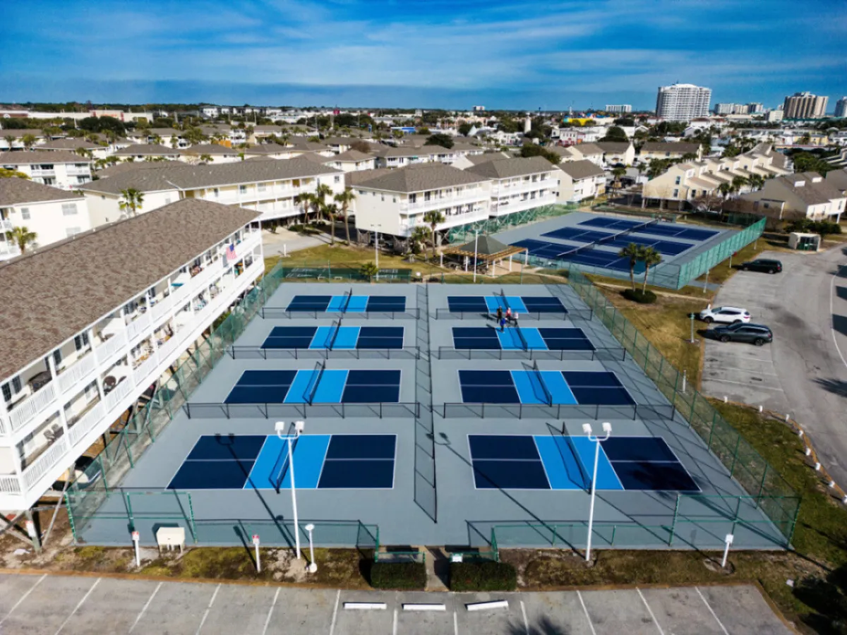 This image captures multiple well-maintained tennis courts with active players engaged in matches. The courts are surrounded by fencing, lighting structures, and nearby residential buildings, conveying a lively and sporty atmosphere.