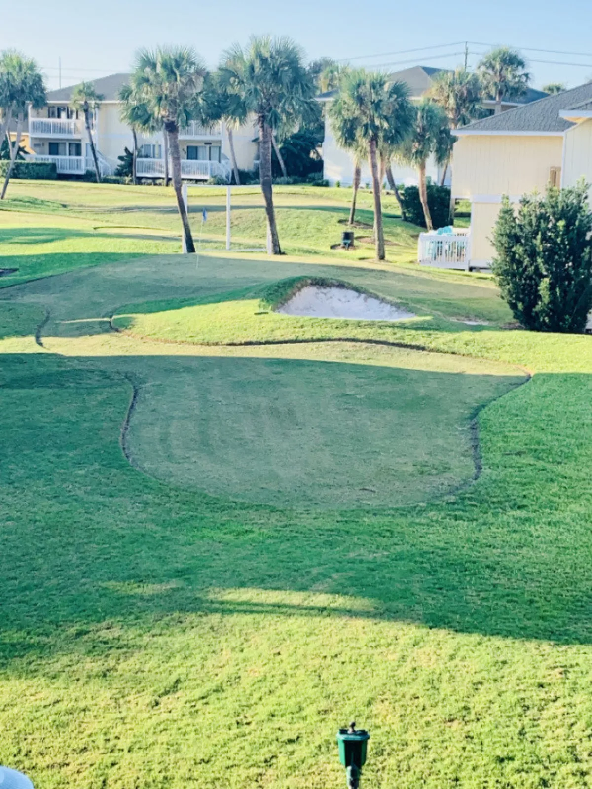A lush golf putting green with a sand bunker is bordered by palm trees and light-colored residential buildings. The setting is tranquil and inviting, with open lawns and clear skies, hinting at the array of recreational options available on-site.