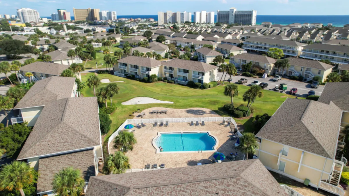 A central swimming pool surrounded by lounge chairs sits amid low-rise condominium buildings and manicured green spaces with palm trees. In the distance, glimpses of the beach and ocean are visible. The scene suggests a vibrant, communal resort atmosphere with amenities like pools, BBQ areas, and proximity to the beach.