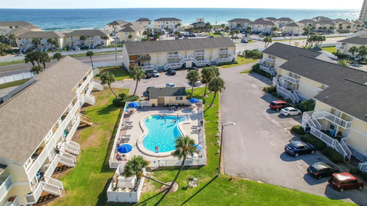 A cheerful scene features a central swimming pool surrounded by lounge chairs and umbrellas, with low-rise condo buildings in the background, well-maintained lawns, palm trees, and a distant view of the beach and ocean. The image radiates a summer vacation vibe and showcases multiple resort amenities.