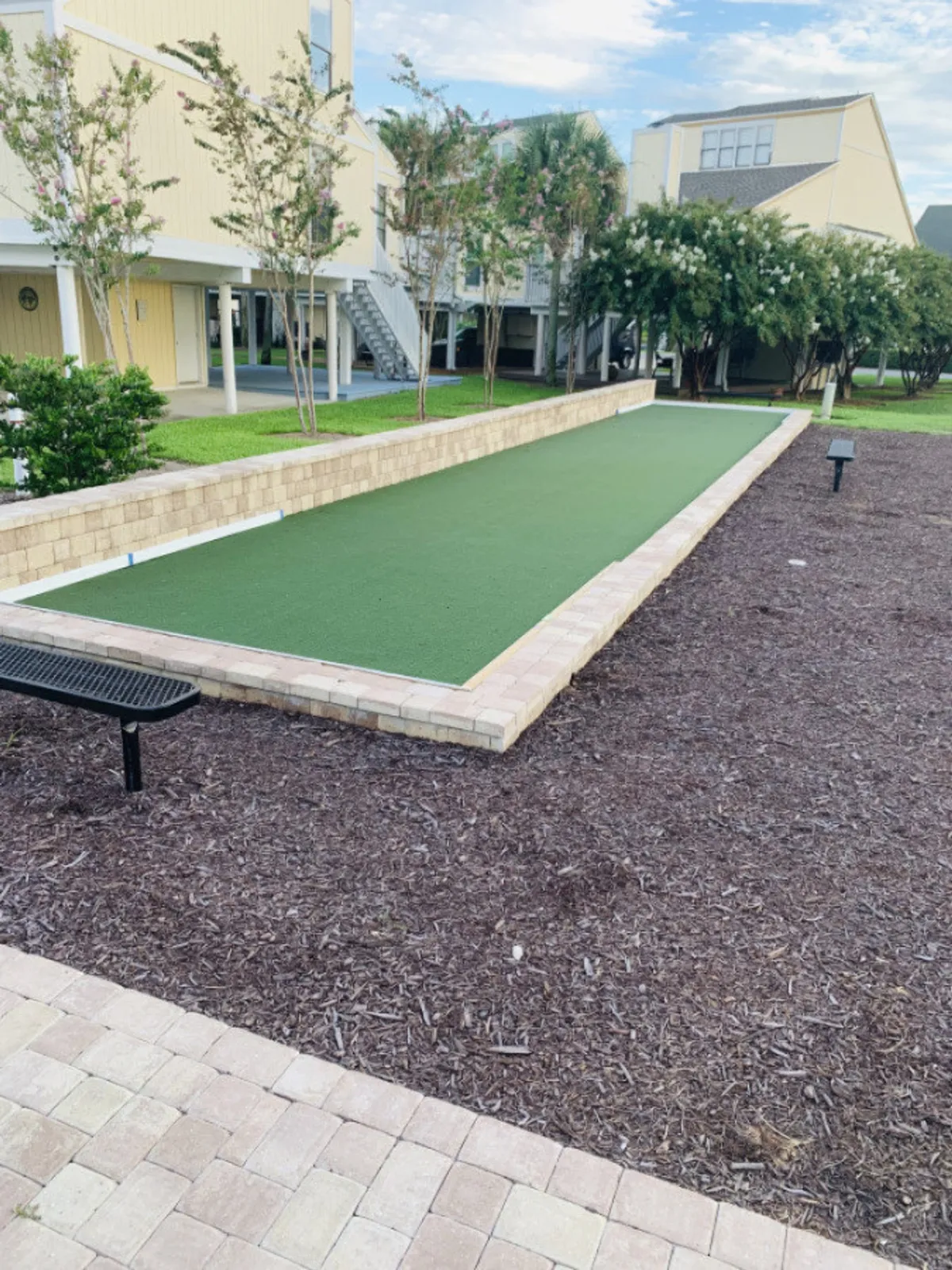 This image features a bocce ball court set amid well-manicured trees and shrubs, with modern yellow coastal buildings in the background and a small black bench nearby. The lush, walkable community grounds are enhanced by a relaxed, social outdoor setting.