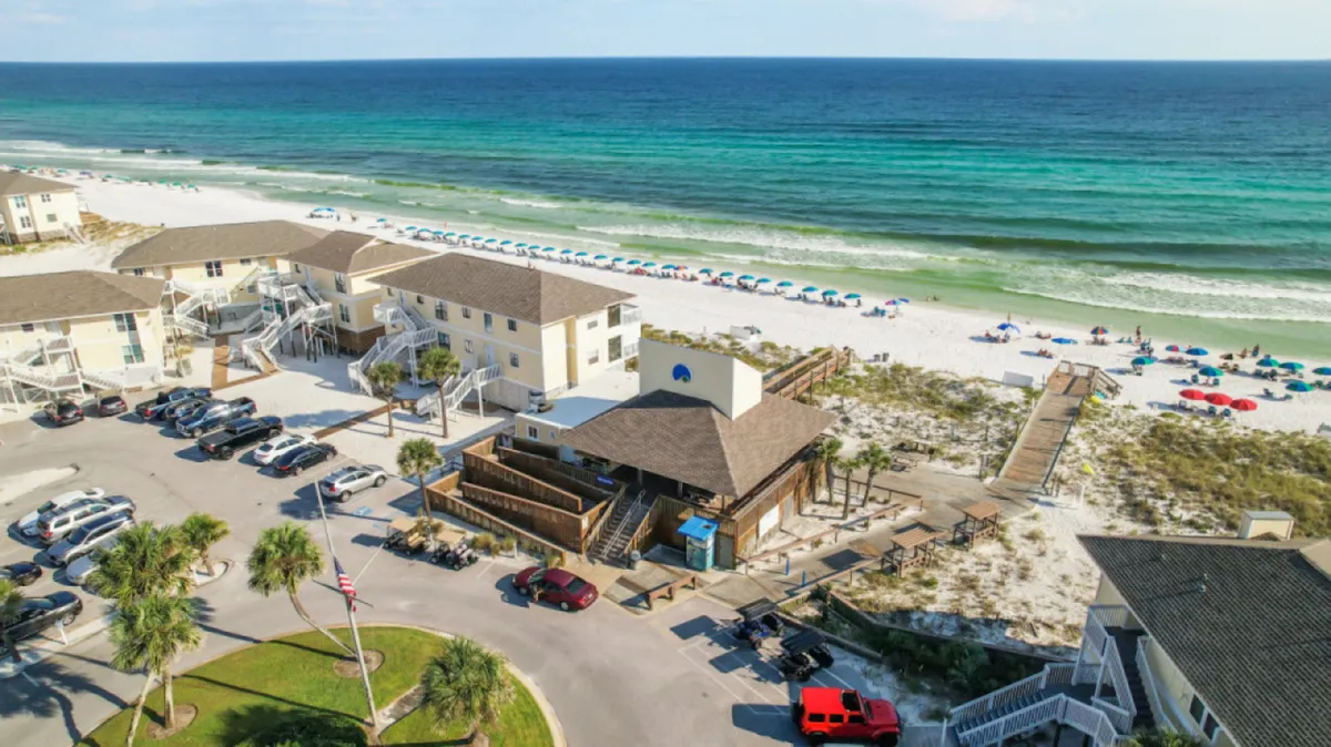 A pristine white sand beach is dotted with colorful umbrellas, with the emerald ocean stretching beyond. Beachfront condo buildings line the shore, and a boardwalk leads directly to the sand, highlighting both recreation and convenient access.