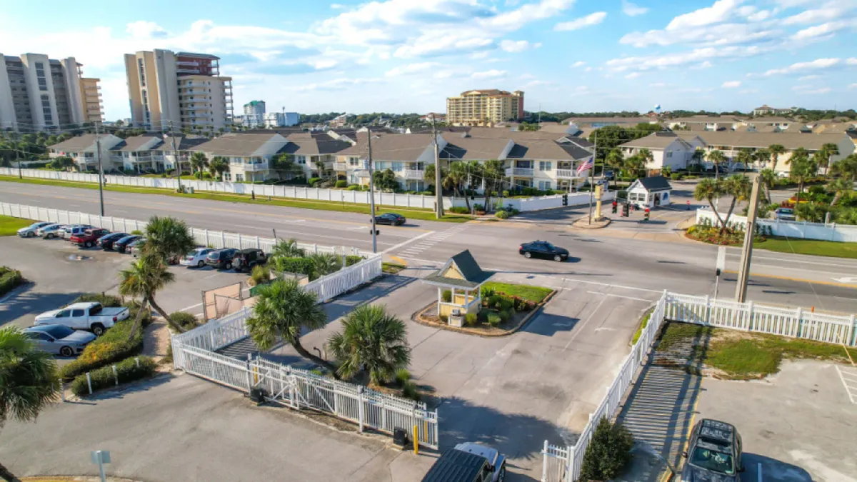 A secure, gated entrance with a security booth, white fencing, palm trees, and adjacent parking lot welcomes residents and guests. Multi-story residential buildings are visible in the background, conveying safety and community.
