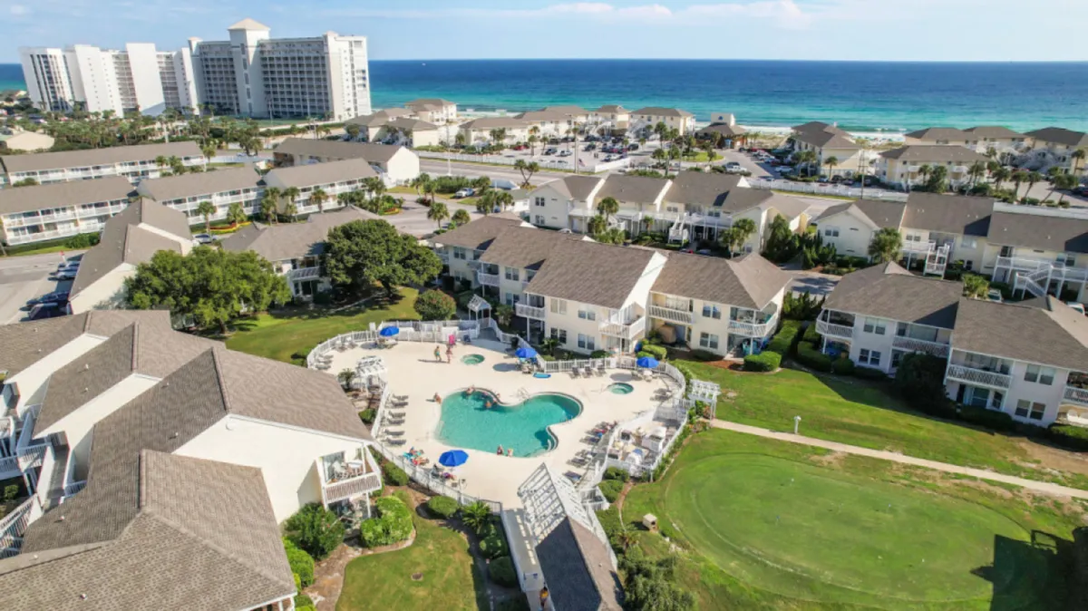 The scene features a central swimming pool with lounge chairs and umbrellas, low-rise condos, an expansive beach and ocean backdrop, and a visible golf course adjacent to the pool. People are swimming, sunbathing, and relaxing, with tennis and golf facilities in view.