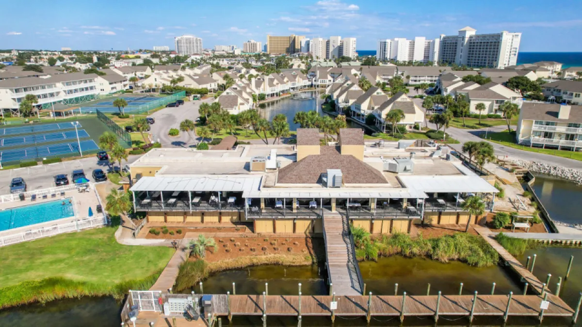 A central clubhouse or restaurant with covered outdoor seating is shown alongside boat docks extending into calm water, a community swimming pool, and multiple tennis courts. The landscaped grounds and water views evoke a sense of leisure and variety.